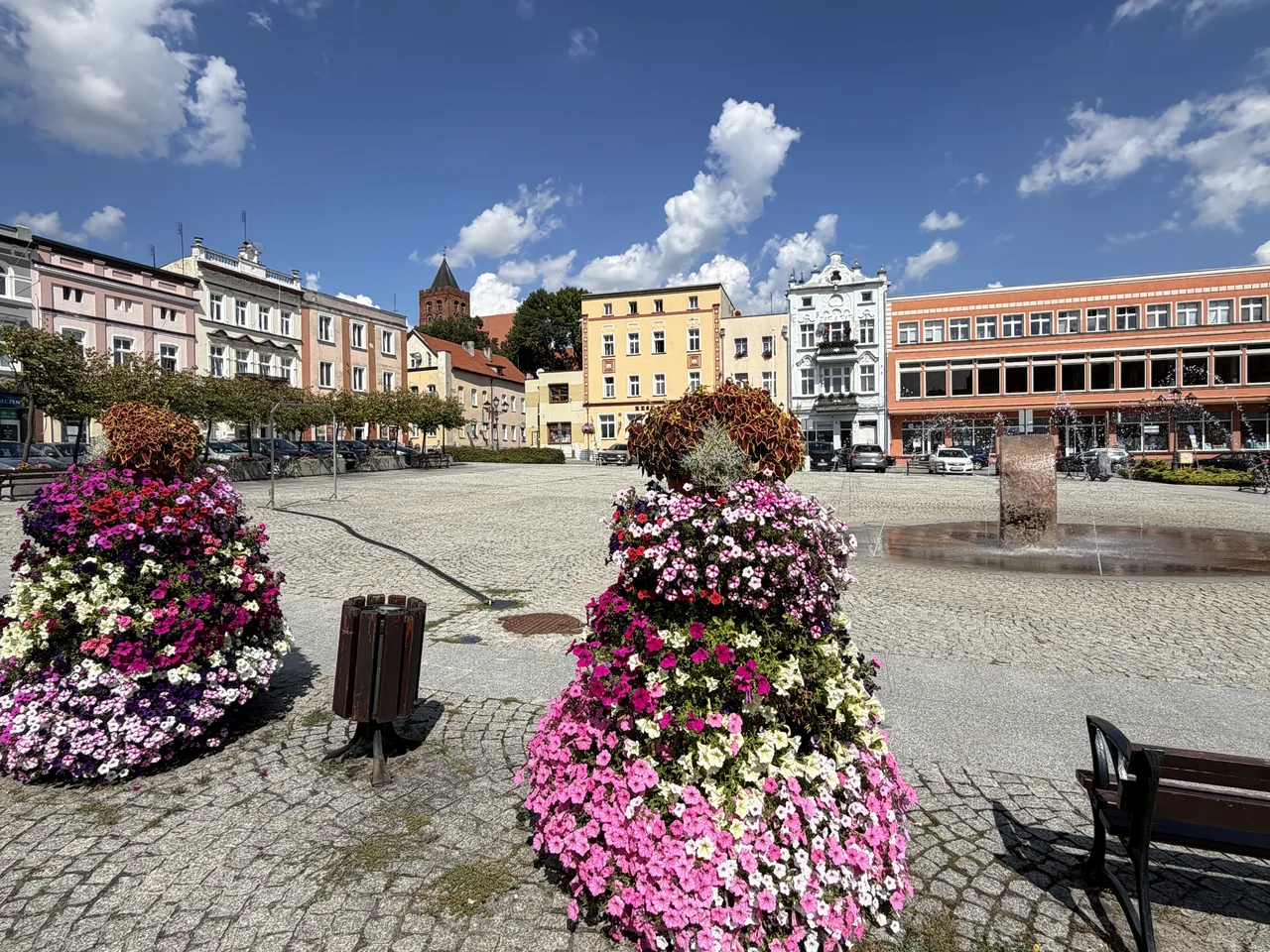 Rynek w Nowem nad Wisłą - kwietniki, fontanna i kamienice, w tle wieża kościoła